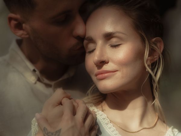 Paar im Profil, sie mit geschlossenen Augen im Sonnenlicht, er küsst sie liebevoll auf die Schlaefe.