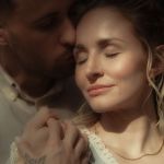 Paar im Profil, sie mit geschlossenen Augen im Sonnenlicht, er küsst sie liebevoll auf die Schlaefe.