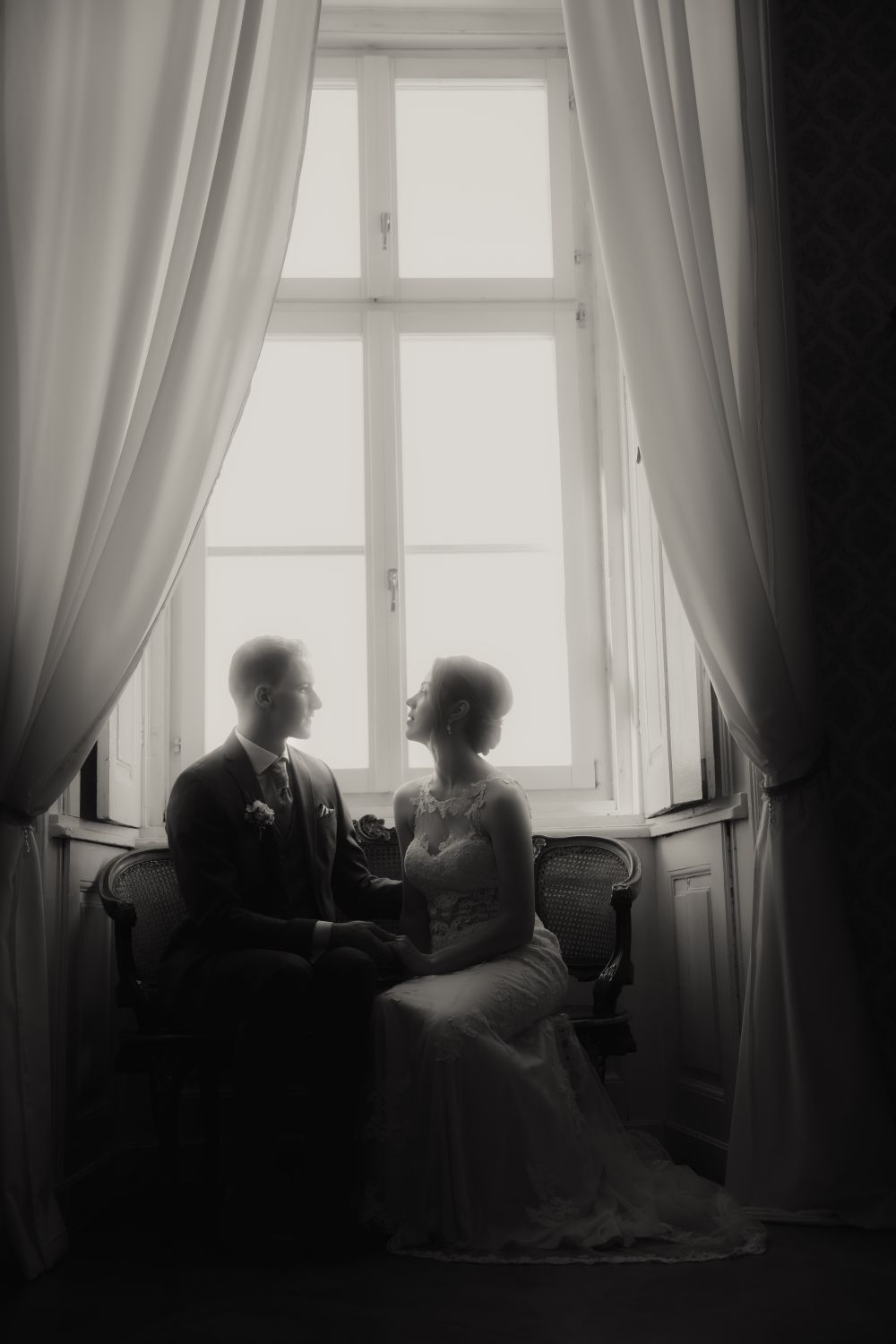 Brautpaar sitzt vor einem Fenster, eingerahmt von langen Vorhängen, blickt sich verliebt an, in schwarz weiß