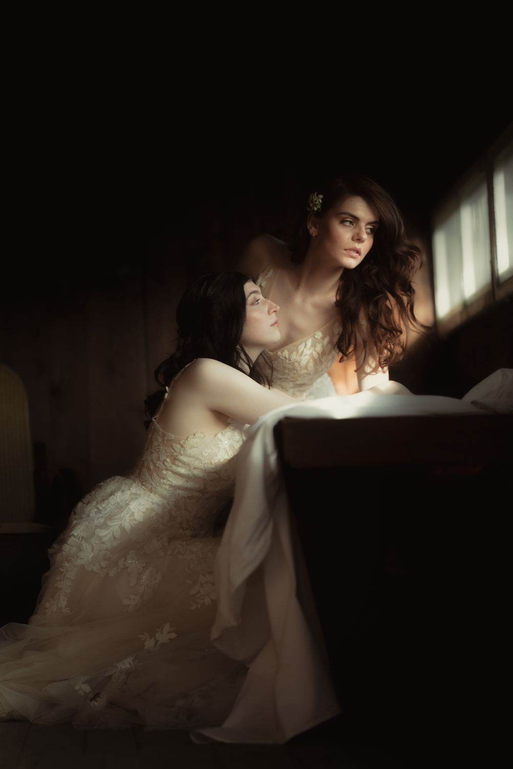 Gleichgeschlechtliches Paar in Spitzenkleidern, in einer dunklen Hütte mit Fenster, eine schaut nachdenklich zum Fensterlicht, die andere Blickt sie an