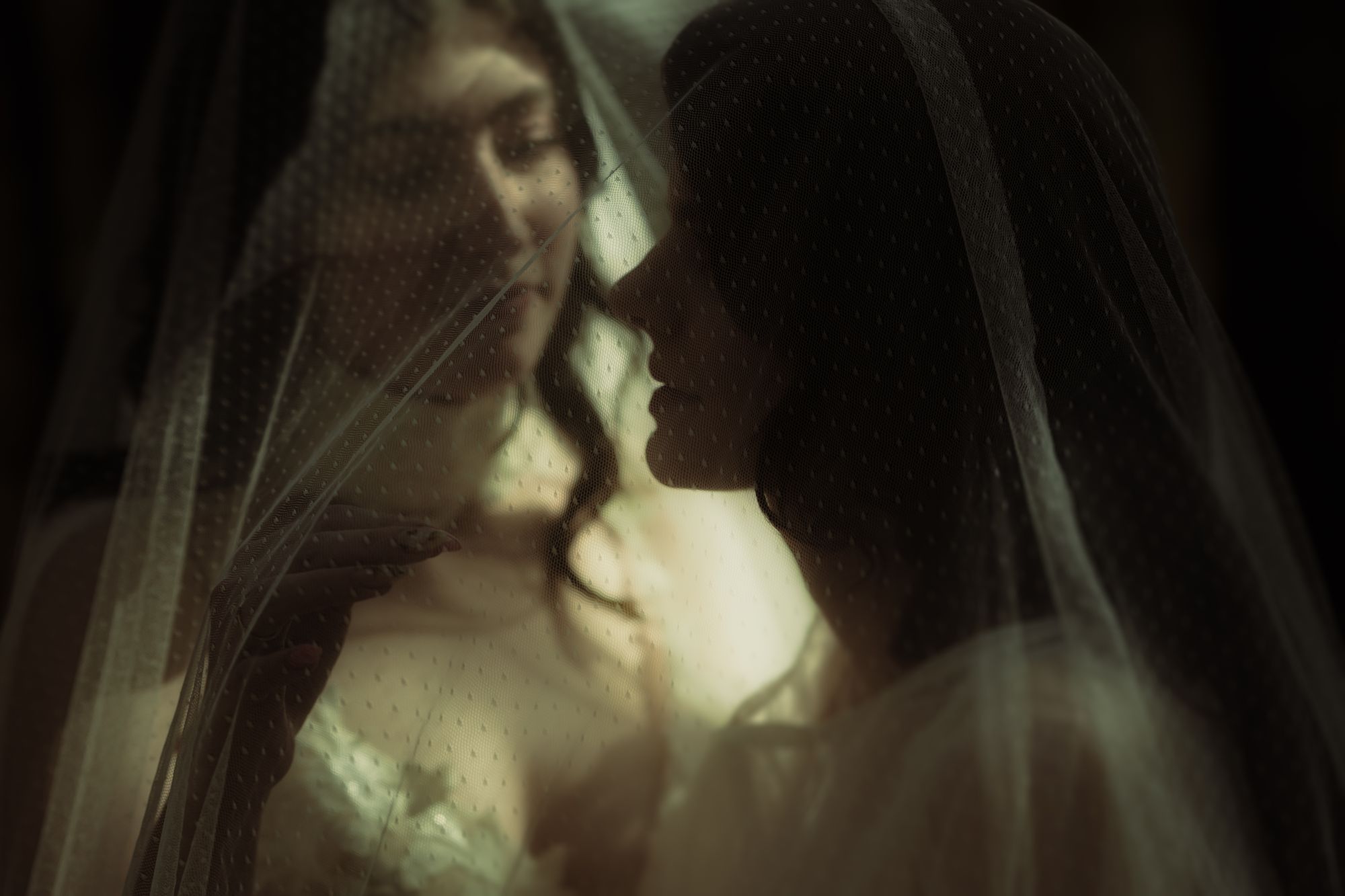 Eine Nahaufnahme von zwei Braeuten, die sich unter einem transparent, gepunkteten Schleier liebevoll anblicken, in weichem Licht