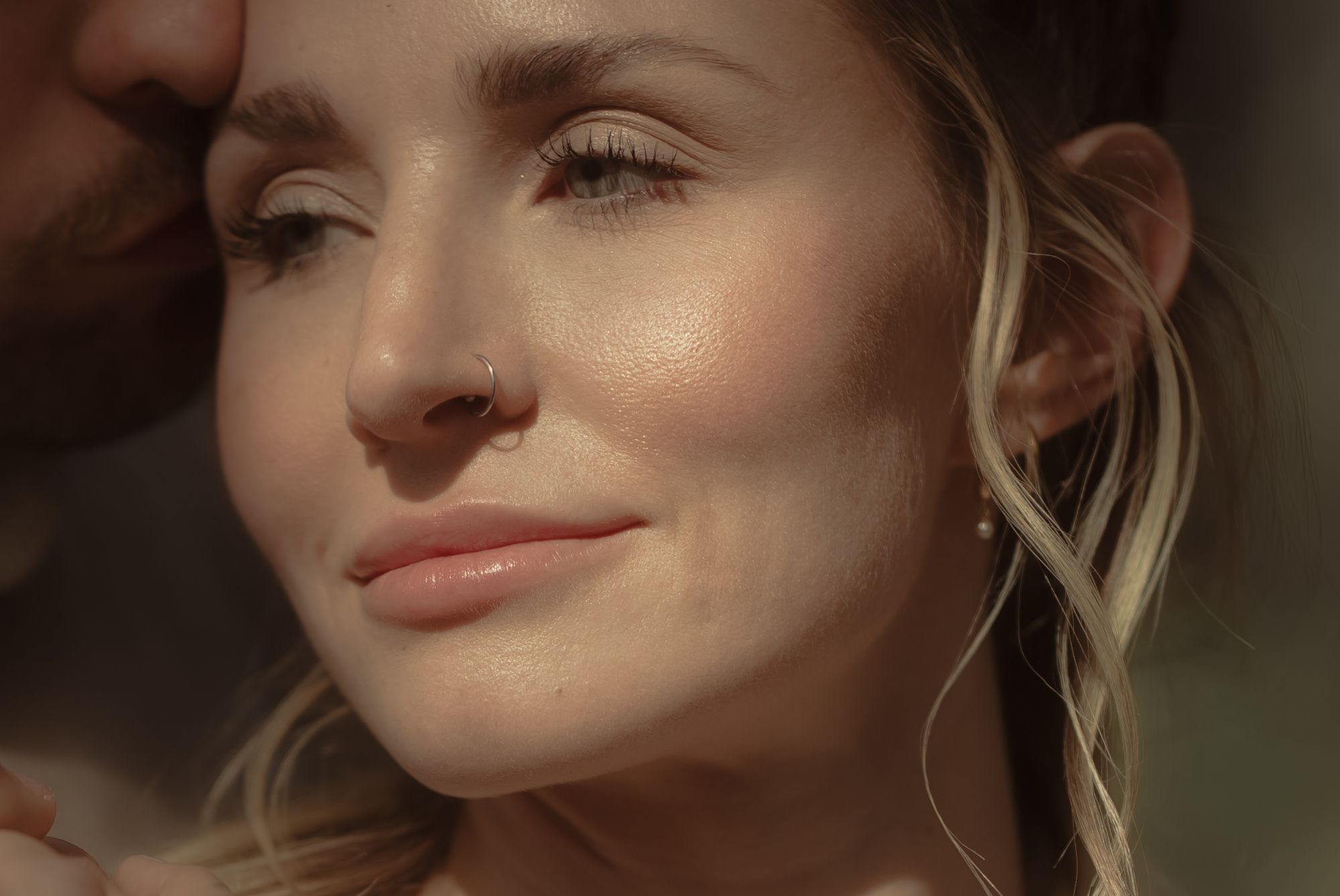 Nahaufnahme einer blonden Frau, die laechelnd in die Ferne blickt, waehrend ihr Partner sie auf die Schlaefe kuesst