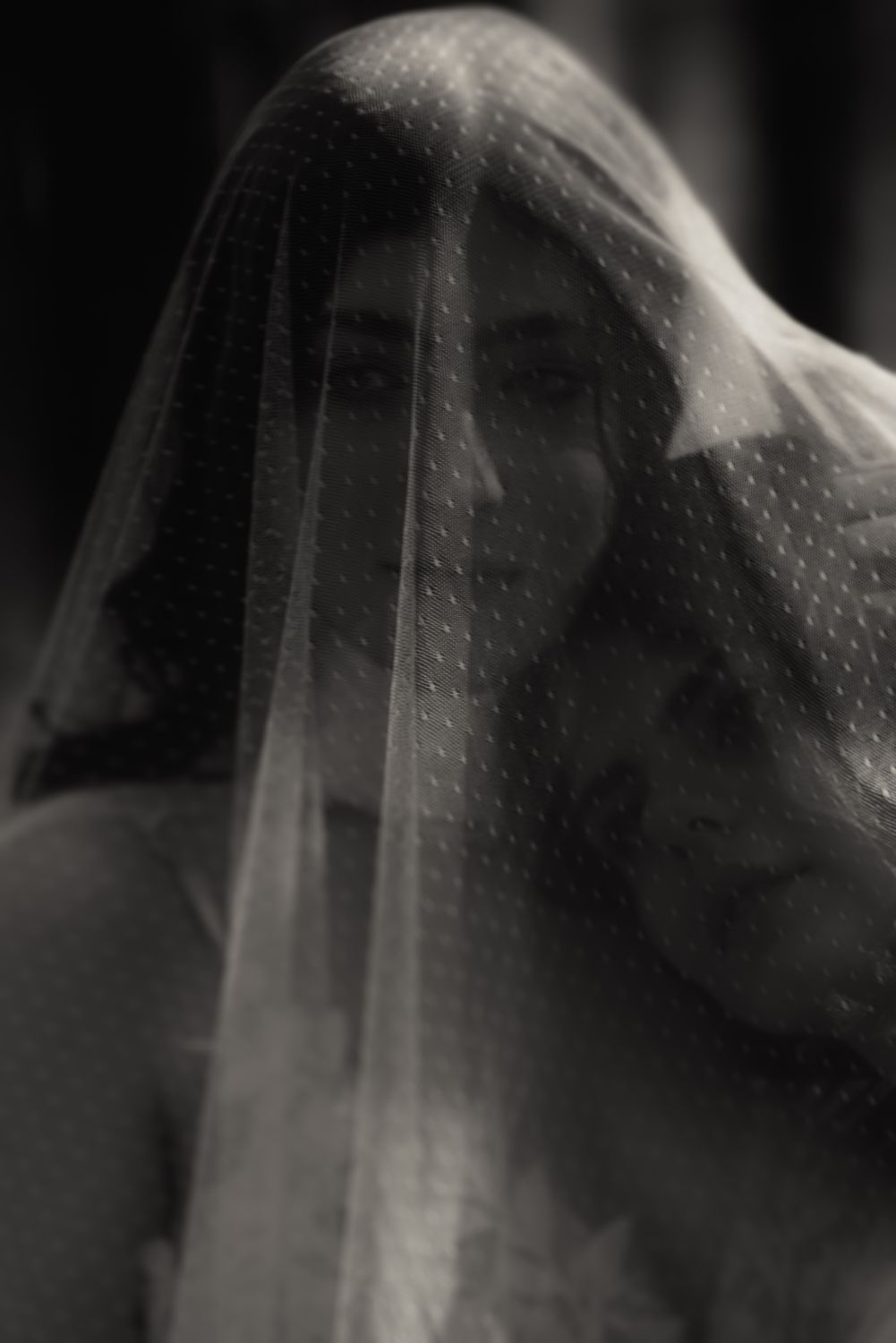 Zwei Braeute in Hochzeitskleidern stehen eng umschlungen unter einem zarten, gepunkteten Schleier, im vertraeumten schwarz weiß Stil